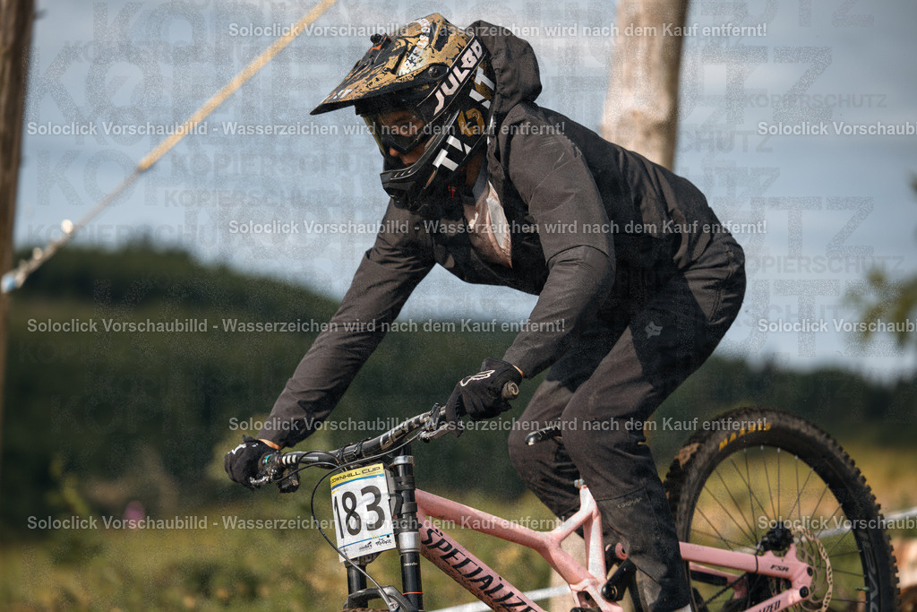 Mountainbiker in Aktion | Ein Mountainbiker in voller Montainbike-Ausrüstung fährt einen staubigen Pfad hinunter. Er trägt einen schwarzen Helm mit dem Schriftzug 'Troy Lee Designs' und eine schwarze Jacke mit dem Logo 'Fox Racing'. Er trägt eine Nummer 83 auf seinem Rücken. Das Mountainbike hat einen pinkfarbenen Rahmen und schwarze Reifen. Im Hintergrund ist eine grüne Landschaft mit Bäumen und Büschen zu sehen. - Realisiert mit Pictrs.com