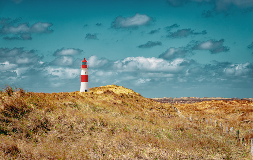 Leuchtturm List-Ost-Sylt-Ellenbogen-umrahmt von Strandhafer | Der Leuchtturm List-Ost befindet sich auf der Halbinsel Ellenbogen im Norden von Sylt.Er wurde 1857 erbaut und ging am 1. Januar 1858 in Betrieb.Der Turm ist rot-weiß geringelt und seit 1977 ferngesteuert.Die Umgebung ist bekannt für ihre weite Dünenlandschaft und ist ein beliebtes Ziel für Wanderungen. - Realisiert mit Pictrs.com