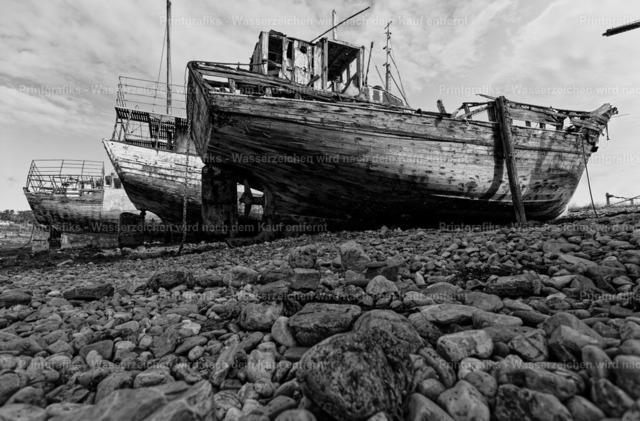 Schiffswrack am steinigen Ufer | Schwarzweißaufnahme eines alten, verwitterten Schiffswracks an einem steinigen Küstenabschnitt. Die niedrige Perspektive, die rauen Holzstrukturen und die Steine im Vordergrund verstärken den dokumentarischen, maritimen Charakter des Motivs. - Realisiert mit Pictrs.com