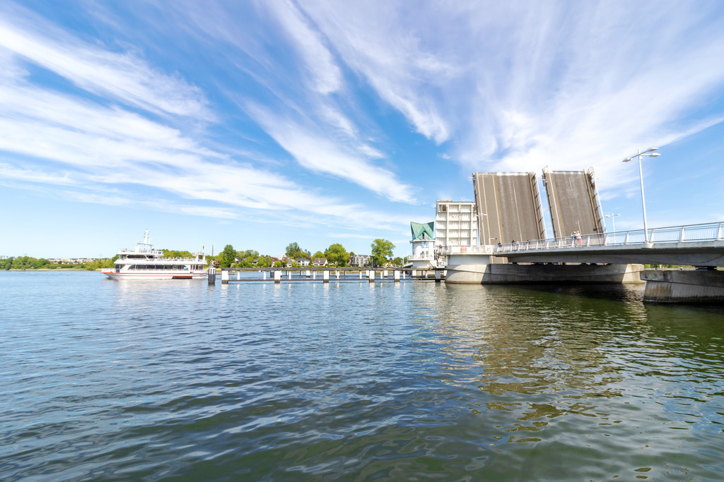 Wandbild: Fähre an geöffneter Schleibrücke | Dieses Wandbild im Querformat zeigt die geöffnete Schleibrücke in Kappeln im Frühling. Auf der Schlei befindet sich die Schleifähre. Am blauen Himmel sind viele Schleierwolken zu sehen.  - Realisiert mit Pictrs.com