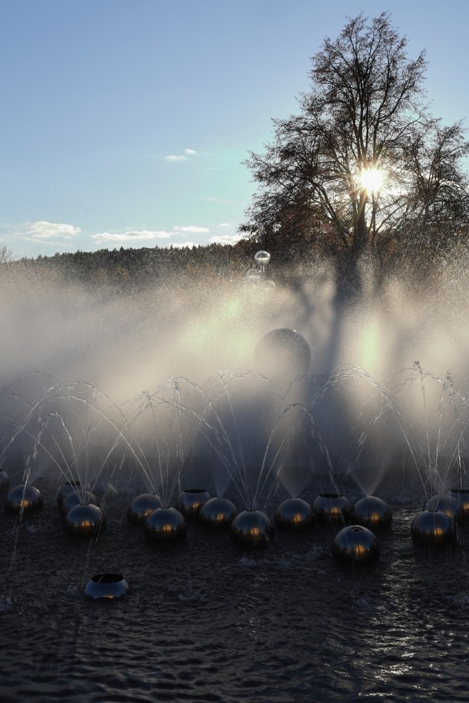 Singende Fontäne Marienbad | Das Foto zeigt die sprudelnde Singende Fontäne in Marienbad (Mariánské Lázně), deren Wasserstrahlen im Sonnenlicht eindrucksvoll aufleuchten. Die Fontäne bildet einen lebendigen Mittelpunkt der Aufnahme, während das Wasser in zahlreichen Bögen und feinen Tropfen emporsteigt und anschließend glitzernd in das Becken zurückfällt. Das Sonnenlicht lässt die Wassertropfen wie kleine Lichtpunkte funkeln und hebt die Bewegung des Wassers besonders schön hervor. Rund um die Fontäne laden die gepflegten Parkanlagen und die historische Architektur des Kurortes zum Verweilen ein. Die Verbindung aus sprudelndem Wasser, Licht und der eleganten Umgebung vermittelt die typische Atmosphäre eines traditionsreichen böhmischen Kurortes. Die Aufnahme wirkt lebendig, heiter und zugleich festlich. Besonders reizvoll ist das Zusammenspiel von Wasser und Sonnenlicht, das der berühmten Marienbader Fontäne eine fast märchenhafte Ausstrahlung verleiht. - Realisiert mit Pictrs.com