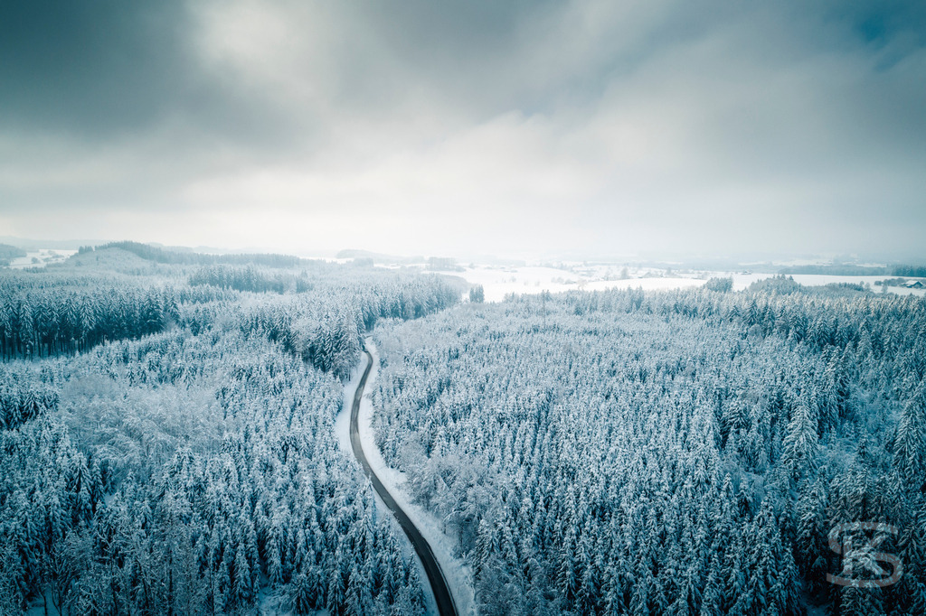 Winterlandschaft Oberschwaben – Verschneiter Wald und Straße aus der Luft | Diese Drohnenaufnahme zeigt eine weitläufige Winterlandschaft in Oberschwaben. Eine geschwungene Straße führt durch dichten, schneebedeckten Nadelwald und öffnet sich zu verschneiten Feldern. Die Luftperspektive offenbart die sanften Hügel und die klare Struktur der Landschaft unter bewölktem Winterhimmel. - Realisiert mit Pictrs.com
