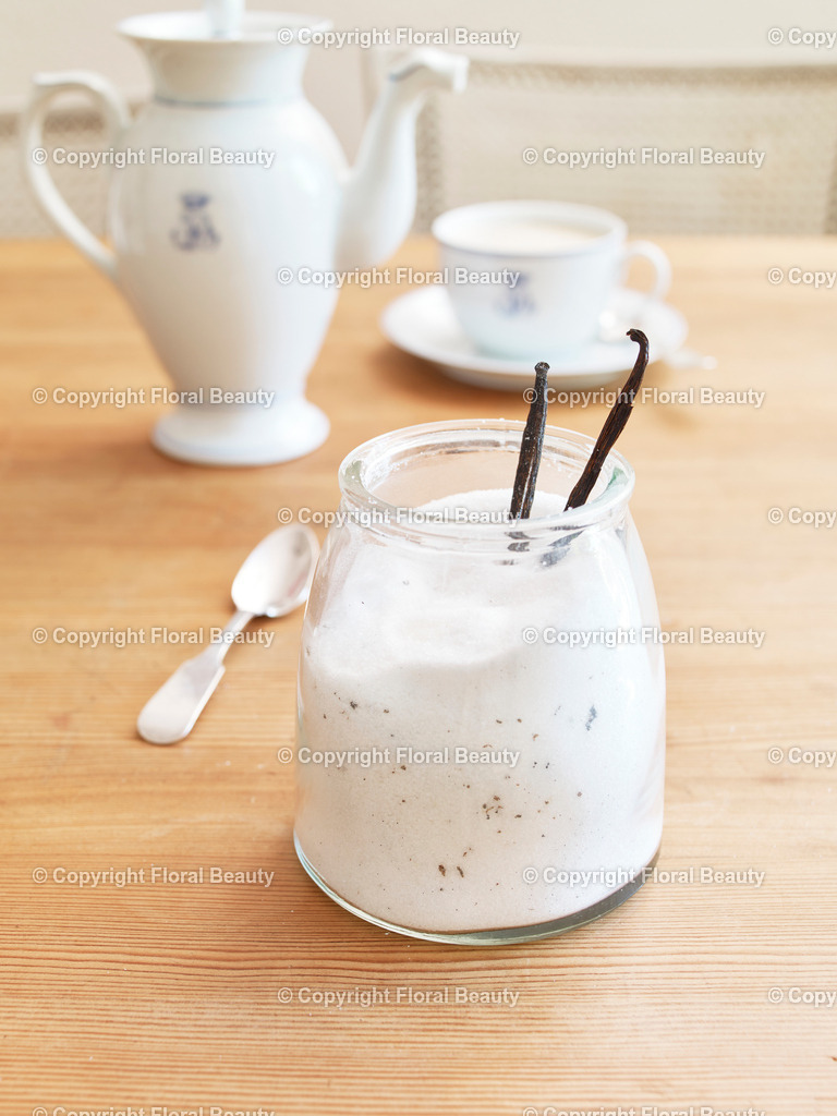 Vanillezucker | Glas gefüllt mit Zucker und Vanilleschoten auf Tisch mit Kaffeegedeck im Hintergrund. - Realisiert mit Pictrs.com