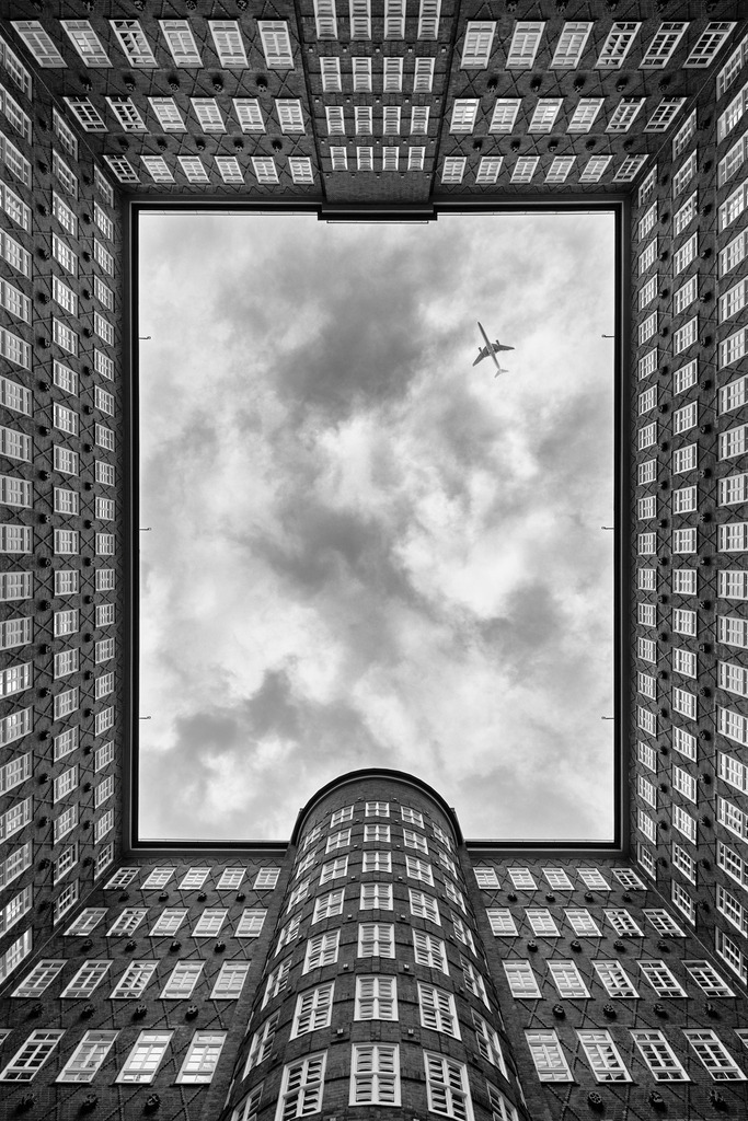 10250601 - Sprinkenhof Weitblick | Die strenge Geometrie des Backsteinbaus trifft auf den offenen Himmel.Mitten im Kontorhausviertel eröffnet sich eine Perspektive, die kaum jemand wahrnimmt – und gerade deshalb fasziniert.Der Himmel wird Teil der Komposition – durchbrochen von einem Flugzeug, das den Blick lenkt.