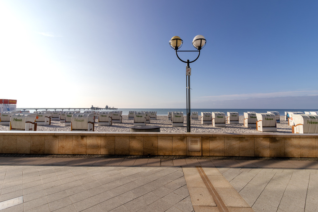 Wandbild: Geordnete Ruhe – Strandpromenade mit Blick aufs Meer | Dieses Wandbild zeigt eine gepflegte Strandszene mit Blick auf eine Reihe von Strandkörben, die ordentlich auf dem Sand angeordnet sind. Im Vordergrund verläuft eine gepflasterte Promenade, die durch eine niedrige Steinmauer vom Strand getrennt ist. Eine zweiflammige Straßenlaterne steht zentral im Bild und fügt sich harmonisch in die klare Gestaltung der Szene ein. Der Himmel ist wolkenlos und strahlt in klarem Blau, während das Meer ruhig bis zum Horizont reicht. Die Szene wirkt strukturiert und zugleich entspannt – ein Moment der Ordnung und Küstenruhe. Dieses Motiv eignet sich ideal als Wandbild für maritime Wohnkonzepte – ob als Leinwandbild, Acrylglasbild, Alu-Dibond FineArt Print oder als Akustikbild. Ein stilvoller Akzent für Wohnzimmer, Büro oder Ferienwohnung. - Realisiert mit Pictrs.com