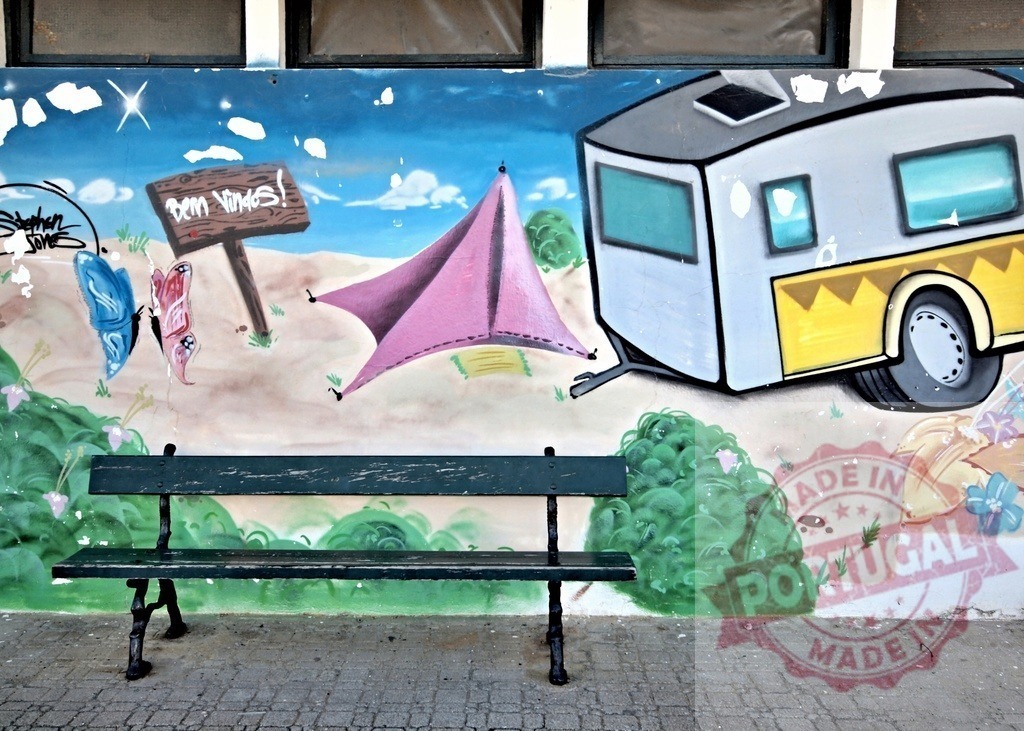 Bench sitting in front of colorful graffiti art depicting camping scene with caravan, tent and butterflies | Empty bench placed near wall with graffiti art showing camping scenery, including caravan, tent, butterflies and bem vindos welcome sign - Realisiert mit Pictrs.com