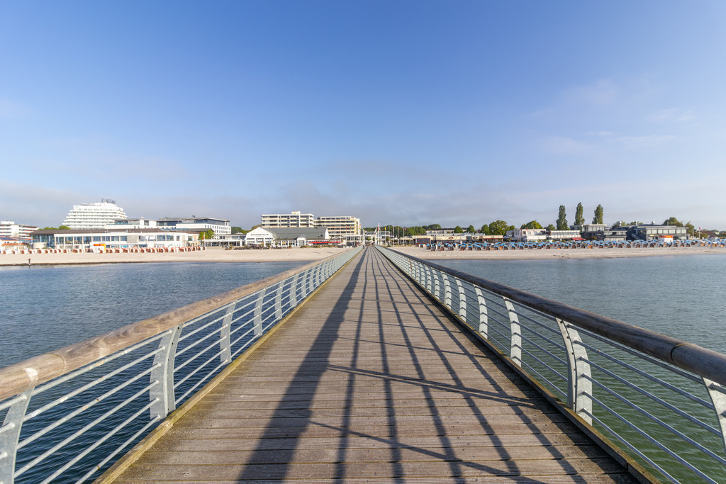 Wandbild: Grömitz Seebrücke – Blick auf den Strand | Dieses maritime Wandbild zeigt den geradlinigen Blick entlang der Seebrücke von Grömitz – eine Perspektive, die Ruhe und Weite vermittelt. Die Holzplanken der Brücke führen den Blick direkt auf den Strand, wo bunte Strandkörbe aufgereiht stehen und die Küstenkulisse prägen. Rechts und links wird die Brücke von Metallgeländern eingefasst, die die klare Linienführung unterstreichen. Im Hintergrund erheben sich moderne Gebäude, darunter Hotels und Ferienanlagen, eingebettet in grüne Baumgruppen. Der Himmel ist leicht bewölkt und verleiht dem Motiv eine freundliche, offene Stimmung. Dieses Bild bringt die entspannte Eleganz eines Ostseetages direkt an die Wand und eignet sich ideal für maritime Wohnkonzepte. Erhältlich als Leinwandbild, Acrylglasbild, Alu-Dibond FineArt Print oder als Akustikbild – ein stilvoller Blickfang für Zuhause, Büro oder Ferienwohnung. - Realisiert mit Pictrs.com