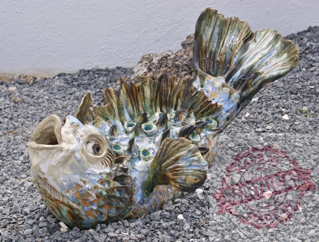Ceramic fish sculpture with vibrant glaze on gravel | Ceramic sculpture of a fish with colorful glaze resting on an outdoor gravel surface - Realisiert mit Pictrs.com