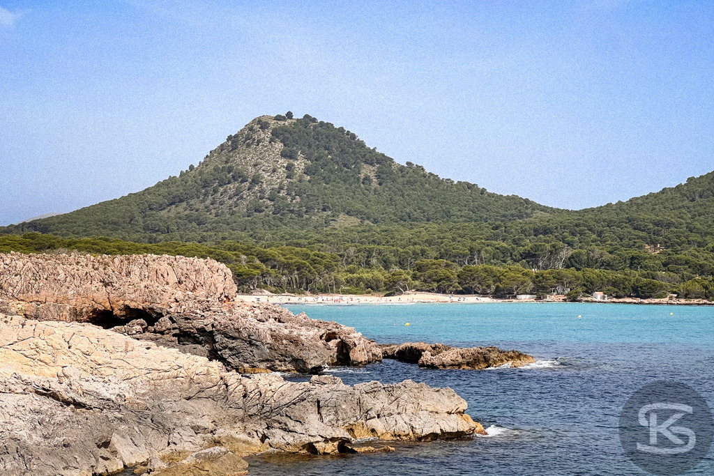 Blick über Felsen auf die Bucht der Cala Agulla und den Berg Es Telégraf, Mallorca | Vordergrund mit hellen Küstenfelsen, dahinter die türkisfarbene Bucht Cala Agulla und ein bewaldeter, kegelförmiger Berg unter blauem Himmel auf Mallorca. - Realisiert mit Pictrs.com