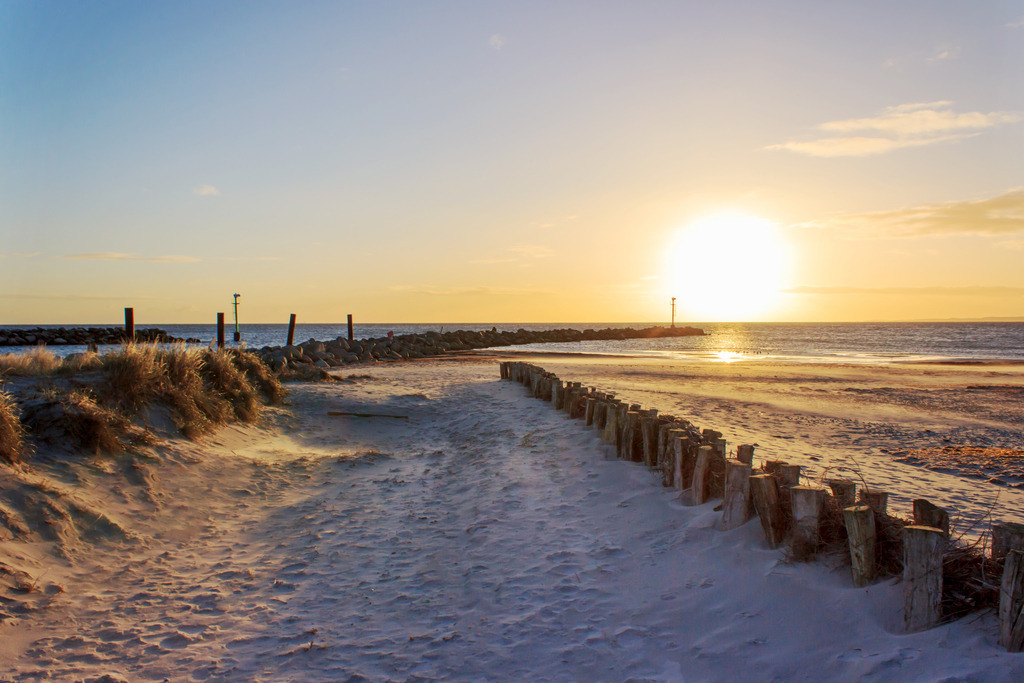 Wandbild: Sonnenaufgang an der Ostsee | Dieses Wandbild im Querformat zeigt einen malerischen Sonnenaufgang am Ostseestrand. Im Vordergrund ist ein Weg ans Meer zu sehen. Auf der rechten Seite befindet sich ein Sandfang und auf der linken Seite wächst Strandhafer vor der Steinmole. - Realisiert mit Pictrs.com