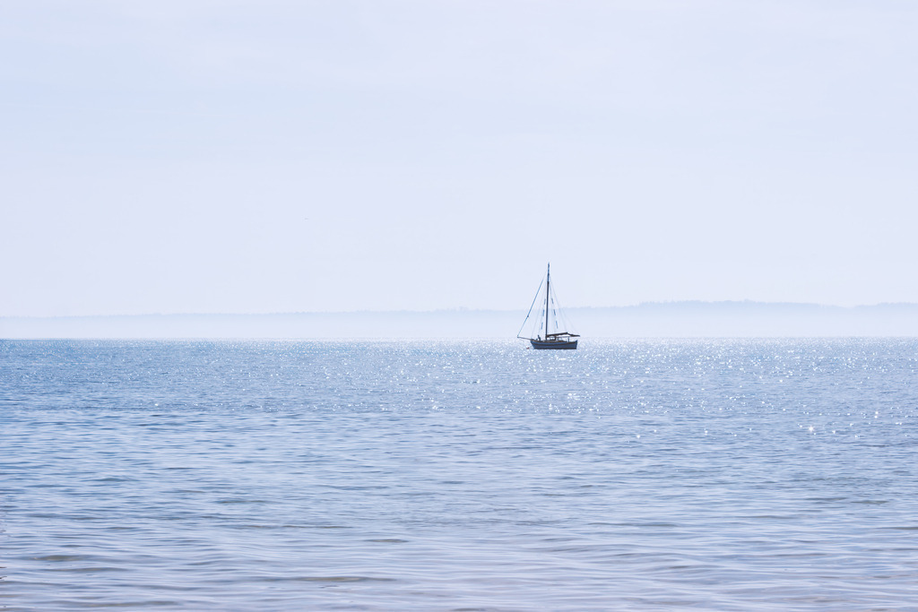 Wandbild: Segelstille auf der Ostsee – Eckernförde | Ein einzelnes Segelboot ruht auf dem weiten Wasser vor Eckernförde, umgeben von hellem Licht und einem klaren Himmel. Die sanften Reflexionen auf der Wasseroberfläche und die minimalistische Komposition verleihen dem Motiv eine friedliche, offene Stimmung. Dieses Bild fängt die maritime Gelassenheit der Ostseeküste ein und vermittelt die besondere Ruhe, die Eckernförde so einzigartig macht.