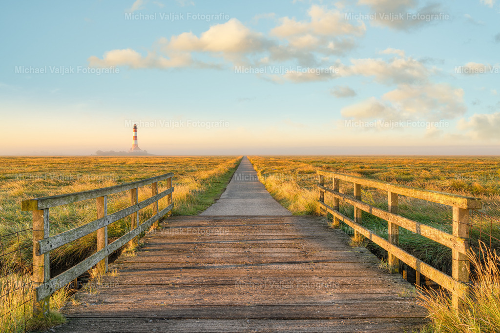 Weg zum Leuchtturm Westerheversand | Kurz nach Sonnenaufgang auf dem Hauptweg zum Leuchtturm Westerheversand. Goldenes Licht streift die Brücke und die Salzwiesen, im Hintergrund liegt der Leuchtturm im leichtem Morgennebel.  - Realisiert mit Pictrs.com