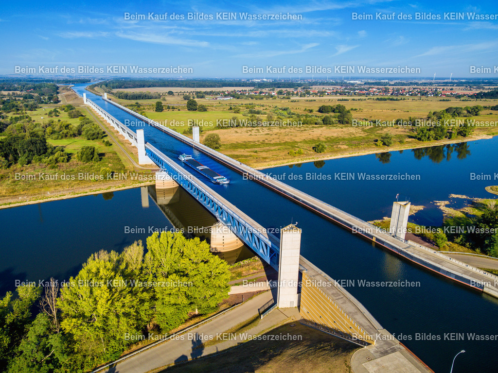 Trogbrücke Wasserstraßenkreuz mit Schiff bei Hohenwarthe-0037 | Das Luftbild zeigt die Trogbrücke des Wasserstraßenkreuzes Magdeburg aus geringer Höhe. Die markante Stahlkonstruktion wird vom intensiven Blau des Wassers und des Himmels geprägt und vermittelt aus der Drohnenperspektive einen eindrucksvollen Eindruck ihrer gewaltigen Dimensionen. Gerade in diesem Moment überquert ein Binnenschiff die Kanalbrücke und macht die besondere Funktion des Bauwerks anschaulich.Die Trogbrücke wurde 2003 in Betrieb genommen und ist mit einer Gesamtlänge von 918 Metern die längste Kanalbrücke Europas. Sie führt den Mittellandkanal über die Elbe und bildet das Herzstück des Wasserstraßenkreuzes Magdeburg. Der wassergefüllte Brückentrog ist 34 Meter breit und rund 4,25 Meter tief. Dadurch können auch große Gütermotorschiffe und Schubverbände mit einer Länge von bis zu 185 Metern die Brücke sicher passieren, ohne den Fluss selbst befahren zu müssen.Die Drohnenaufnahme verdeutlicht eindrucksvoll die ingenieurtechnische Leistung dieses außergewöhnlichen Bauwerks. Das über die Elbe fahrende Schiff unterstreicht die Dimensionen der Trogbrücke und macht sichtbar, wie moderne Wasserbautechnik den europäischen Binnenwasserverkehr effizient und unabhängig von den Wasserständen der Elbe ermöglicht. - Realisiert mit Pictrs.com