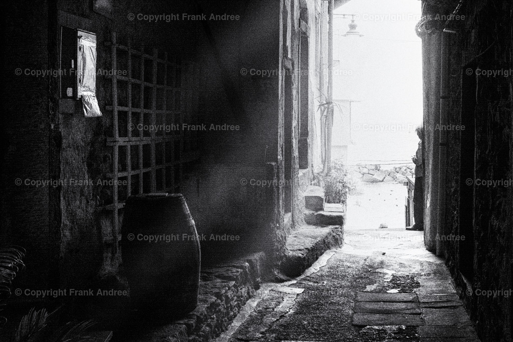 Cinque Terre | Ein Mann steht am ende eines Hausdurchganges. Licht fällt in den dunklen Innenraum, wo ein altes Holzfass steht. Straßenfotografie aus Italien in schwarz und weiß. - Realisiert mit Pictrs.com