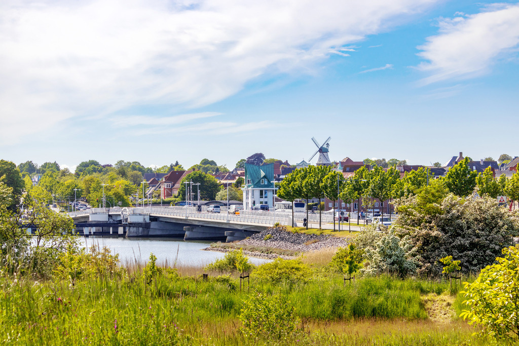 Wandbild: Schleibrücke und Windmühle in Kappeln | Dieses Wandbild im Querformat zeigt die Schleibrücke und die Windmühle in Kappeln im Frühling. Im Vordergrund sind viele frühlingshafte Bäume und Sträucher zu sehen. - Realisiert mit Pictrs.com