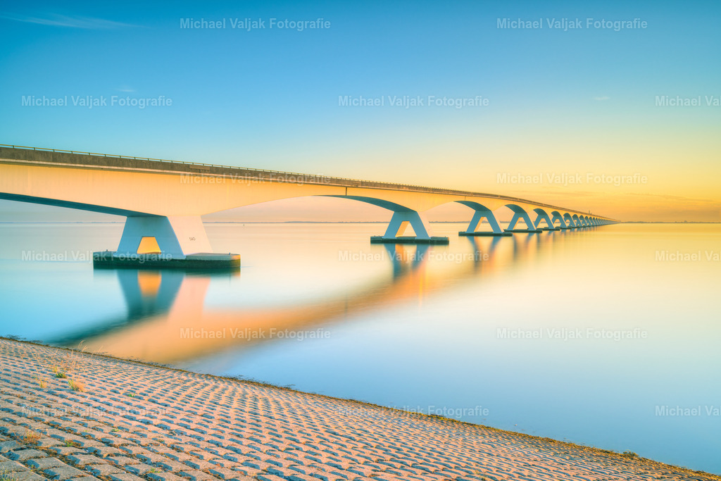 Zeelandbrücke im warmen Morgenlicht | Die ersten Sonnenstrahlen des Tages tauchen die imposante Zeelandbrücke in ein warmes, goldenes Licht. Kurz nach Sonnenaufgang liegt eine besondere Ruhe über der Küstenlandschaft von Zeeland, während sich die lange Reihe der Brückenpfeiler weit über das Wasser erstreckt.Durch die Langzeitbelichtung erhält das Wasser eine weiche, fast samtige Struktur und bildet einen stimmungsvollen Kontrast zu den klaren Linien der Brücke. Das frühe Morgenlicht lässt die Szenerie in warmen Farbtönen erstrahlen und verleiht dem Motiv eine ruhige, zeitlose Atmosphäre.Ein atmosphärisches Landschaftsfoto der Zeelandbrücke in Zeeland, Niederlande, das die besondere Stimmung eines neuen Tages an der niederländischen Küste einfängt. - Realisiert mit Pictrs.com