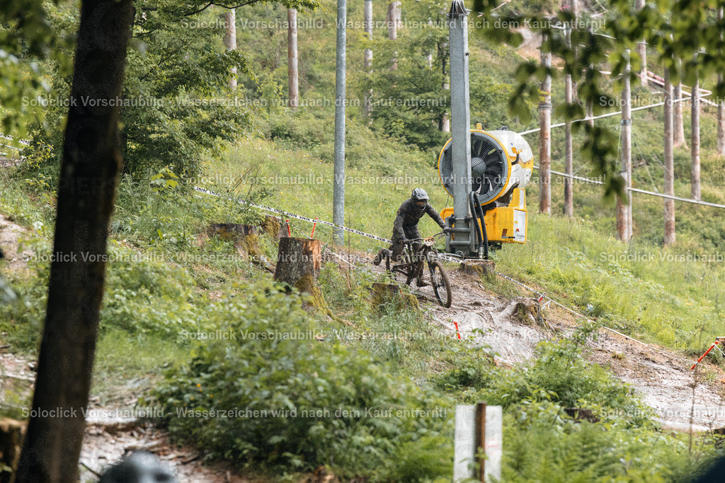 Mountainbike-Abenteuer im Gelände | Das Bild zeigt einen Mountainbiker in voller Ausrüstung, der einen steilen, mit Wurzeln und Schlamm übersäten Hang hinunterfährt. Er trägt einen schwarzen Helm und eine dunkle Jacke. Im Hintergrund steht eine gelbe Schneekanone, die auf einem Stahlgerüst montiert ist. Der Hang ist von dichtem Grün bewachsen, und es gibt mehrere Baumstümpfe im Vordergrund. Das Bild vermittelt ein Gefühl von Geschwindigkeit und Abenteuer. - Realisiert mit Pictrs.com