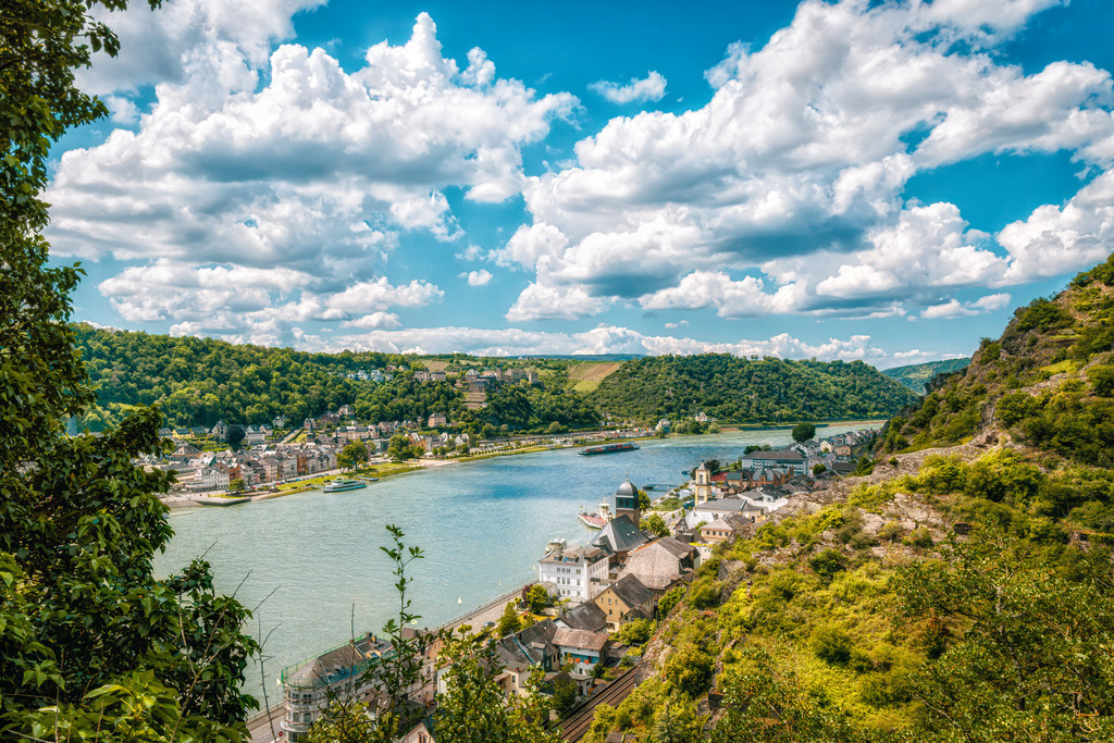 Blick in den Mittelrhein auf Sankt Goar und Sankt Goarshausen | Dieses Bild zeigt das UNESCO-Welterbe Oberes Mittelrheintal in Deutschland, genauer gesagt den Blick auf den Rhein bei Sankt Goarshausen (rechts im Vordergrund) und das gegenüberliegende Sankt Goar (links im Hintergrund).Mittelrheintal: Eine der ältesten und bedeutendsten Kulturlandschaften Europas, weltberühmt für ihre Dichte an mittelalterlichen Burgen und steilen WeinbergenLoreleystadt: Die Region ist eng mit der Sage der Loreley verknüpft, da der berühmte Schieferfelsen nur wenige Kilometer stromaufwärts liegt.Historische Bauwerke: Auf den umliegenden Hügeln thronen markante Festungen wie die Burg Katz über Sankt Goarshausen und die Burg Rheinfels auf der anderen Rheinseite - Realisiert mit Pictrs.com