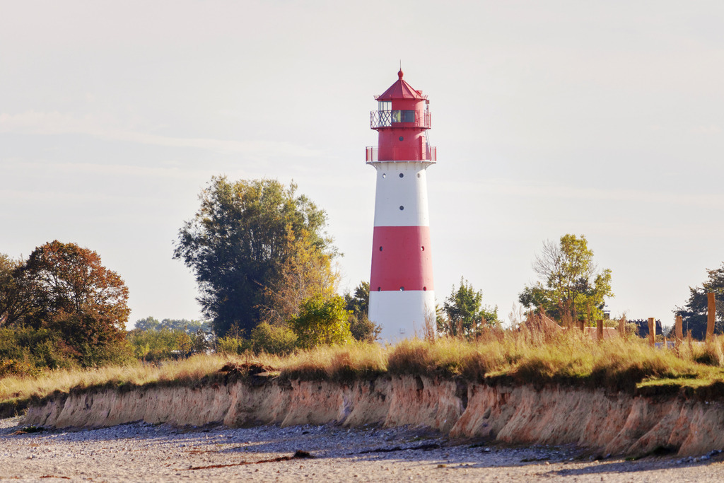Wandbild: Leuchtturm in Falshöft | Dieses Wandbild im Querformat zeigt den Leuchtturm in Falshöft im Herbst. Im Vordergrund ist der Strand sowie eine kleine Steilküste zu sehen. In der Mitte des Bildes ist der Leuchtturm zu sehen. Durch den eingesetzten Telezoom wirkt es fast so als wenn der Leuchtturm am Strand steht.   - Realisiert mit Pictrs.com