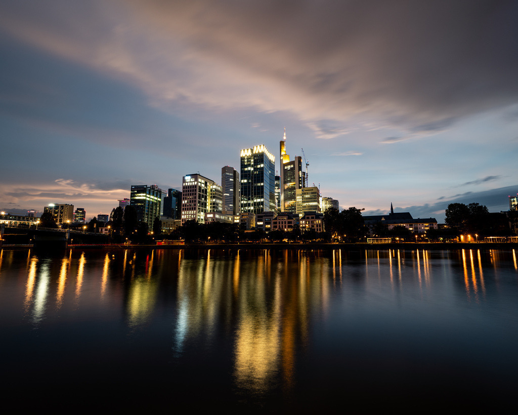 Skyline von Frankfurt am Main zur blauen Stunde | Die beleuchteten Hochhäuser der Frankfurter Skyline spiegeln sich zur blauen Stunde im Main. Das ruhige Wasser und die ziehenden Wolken verleihen der Stadtansicht eine elegante Wirkung. - Realisiert mit Pictrs.com