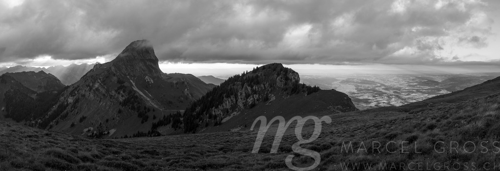 Dramatic Swiss Alps Landscape with Stockhorn and surroundings | A wide panoramic view showcases a rugged mountain range under a dramatic, cloudy sky. The foreground features a grassy slope leading up to the peaks, while a valley stretches out in the distance, partially obscured by the clouds. The scene evokes a sense of natural grandeur and the raw beauty of the Swiss Alps. - Realisiert mit Pictrs.com