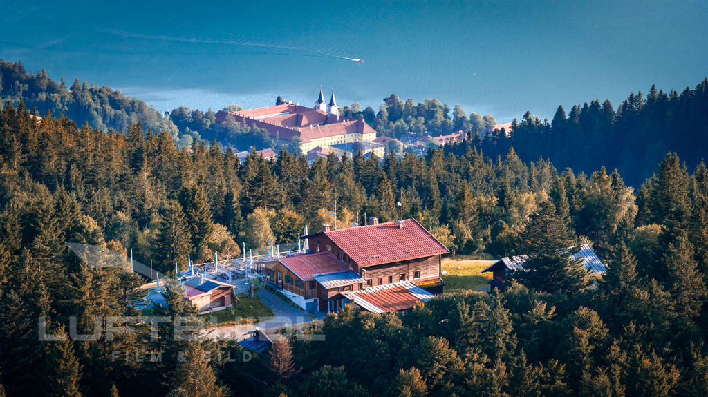 Neureuth mit Schloss-Tegernsee-Kulisse | Im Vordergrund liegt die Neureuth, eine traditionsreiche Berghütte oberhalb von Tegernsee, eingebettet in dichte Wälder. Dahinter öffnet sich der Blick über den See hinweg auf das markante Schloss Tegernsee mit seinen Türmen, das am Ufer thront. Die Kombination aus uriger Berggaststätte und geschichtsträchtigem Bauwerk am Wasser zeigt die ganze Vielfalt des Tegernseer Tals. - Realisiert mit Pictrs.com