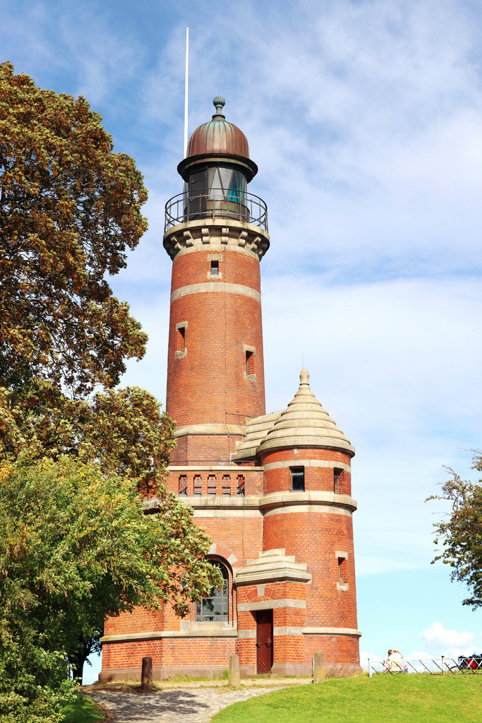 Wandbild: Leuchtturm in Kiel Holtenau | Das Wandbild im Hochformat zeigt den Leuchtturm in Kiel Holtenau im Sommer. Der Leuchtturm an der Einfahrt zum Nord-Ostsee-Kanal befindet sich auf einem kleinen Hügel. Neben dem Leuchtturm stehen einige Bäume. Am blauen Himmel sind einige Wolken zu sehen.  - Realisiert mit Pictrs.com