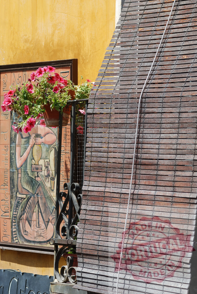 Balcony with flowers, wooden blinds, and street tile art. Cuenca - Spain | Facade showing a balcony with blooming flowers, wooden blinds, and decorative tile art - Realized with Pictrs.com