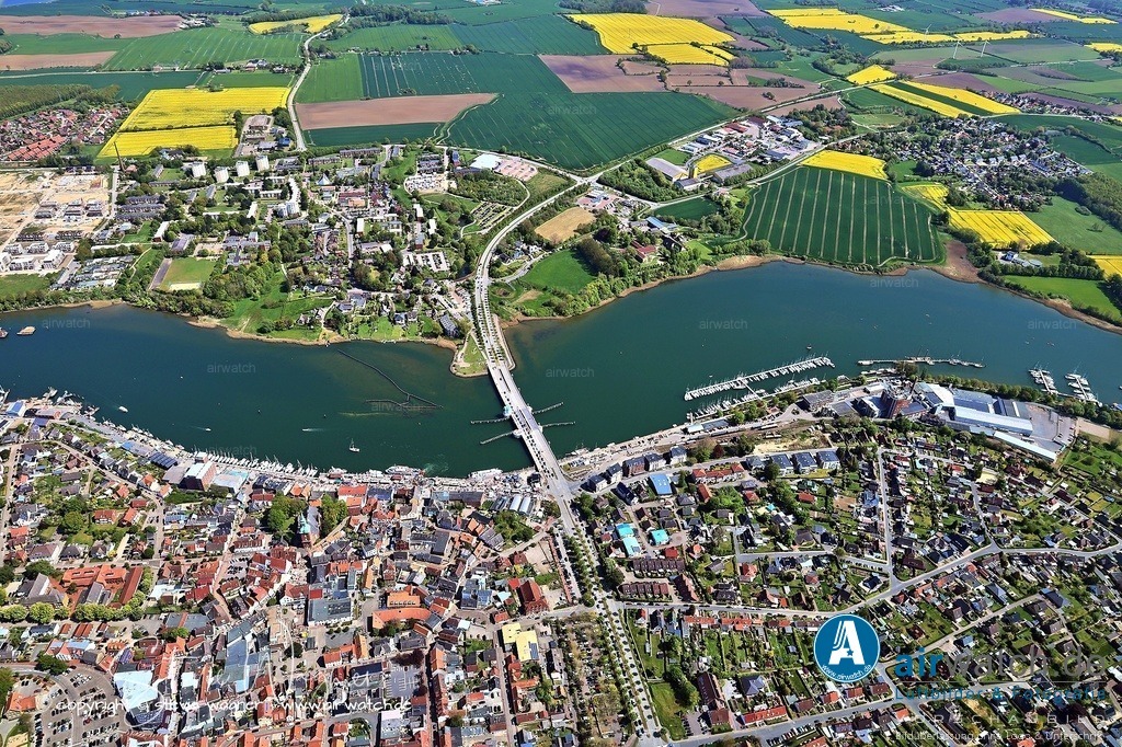 Luftbild Kappeln an der Schlei & Ellenberg | Ein besonderes Erlebnis ist die Spaziergang durch die Innenstadt, bei dem man auf dem Kopfsteinpflaster Fische aus Messing entdecken kann – die sogenannten Heringsplatten, die jeweils eine persönliche Geschichte tragen. Hinter der St. Nikolaikirche befinden sich schattige Holzbänke, von denen aus man einen erhöhten Blick auf die Schlei, den Heringszaun und das Ellenberger Ufer genießen kann. Gleich gegenüber, im Ellenberger Wald, liegt ein ruhiger Strandabschnitt mit einer herrlichen Aussicht auf die Reetdachhäuser in Rabelsund.