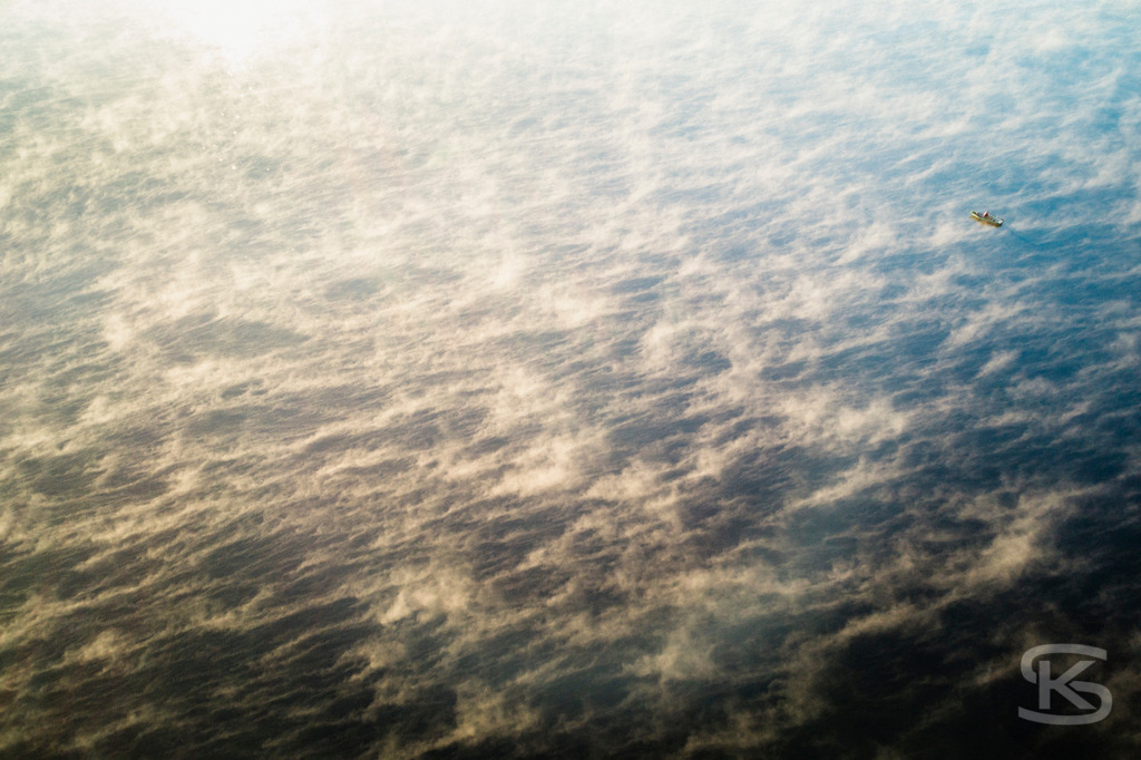 Fischer im Boot auf dem Wasser - Sonnenaufgang, Nebel, Luftaufnahme | Künstlerische Drohnenaufnahme eines einsamen Fischers in seinem Bootr. Minimalistische Vogelperspektive zeigt Nebelschwaden und Lichtreflexionen während des Sonnenaufgangs über der Wasseroberfläche. - Realisiert mit Pictrs.com