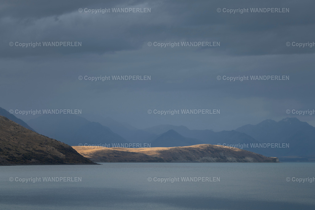 Lake Tekapo |  Eine dramatische und atmosphärische Szene am See von Tekapo. Dunkle, bedrohliche Wolken hängen über der ikonischen Bergkette. Die Oberfläche des Sees bleibt ruhig und glatt. Die Hügel im Vordergrund sind in ein warmes, goldenes Licht getaucht, was einen auffälligen Kontrast zu den kühlen, gedämpften Tönen der entfernten Gipfel und des bewölkten Himmels schafft. Dieses Bild unterstreicht die mächtige und ständig wechselnde Natur der Landschaft der Südinsel Neuseelands. - Realisiert mit Pictrs.com