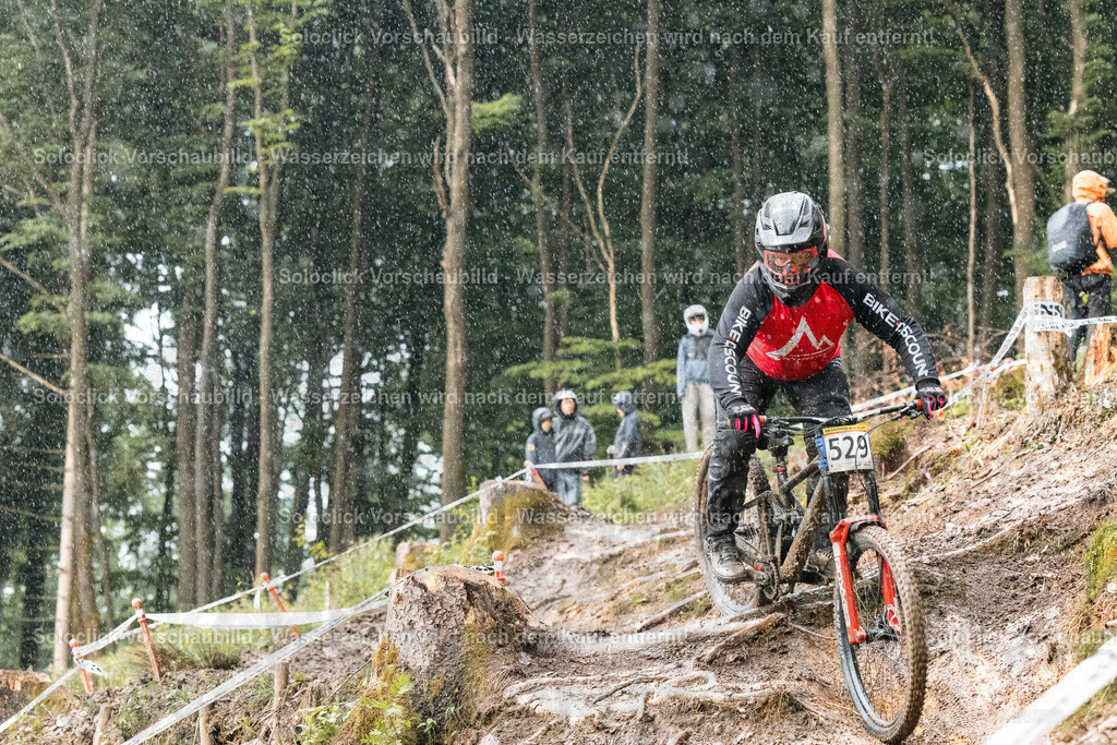 Mountainbiker im Regen | Ein Mountainbiker rast durch ein bewaldetes Gelände, während es regnet. Er trägt einen roten Overall mit dem Schriftzug 'BIKE&amp;COUNT' auf dem Rücken, einen schwarzen Helm und Schutzbrille. Er fährt ein rotes Mountainbike mit schwarzem Rahmen und gelben Akzenten. Im Hintergrund sind weitere Mountainbiker und ein bewaldetes Tal zu sehen. Die Strecke ist mit roten und weißen Markierungen versehen. - Realisiert mit Pictrs.com