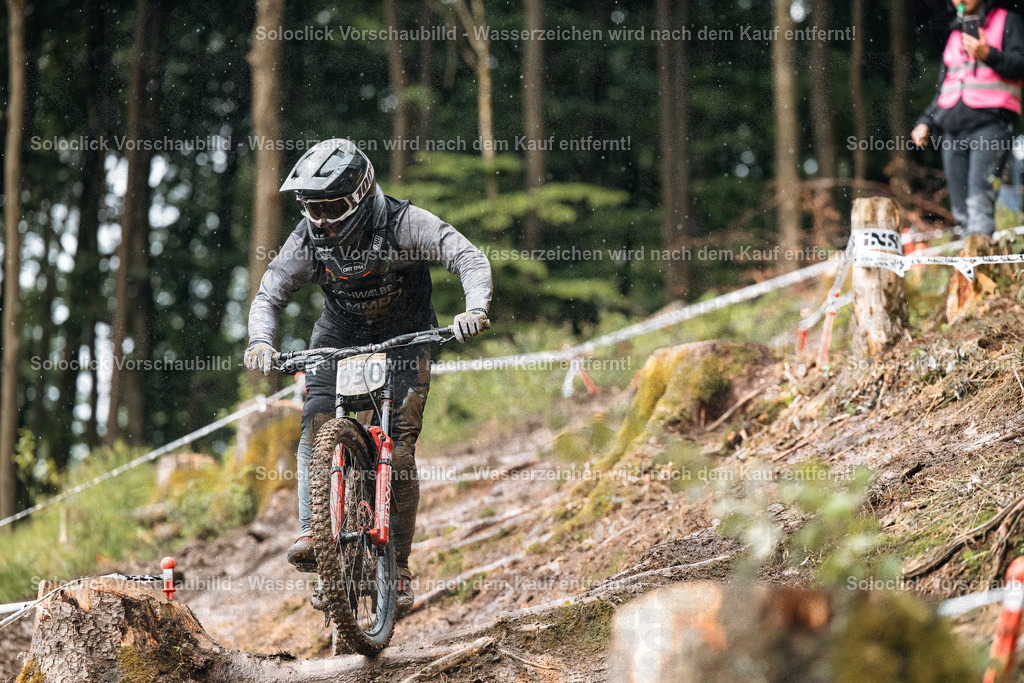 Mountainbiker im Wald | Ein Mountainbiker in voller Ausrüstung rast einen steilen, kurvigen Trail in einem dichten Wald hinunter. Er trägt einen schwarzen Helm mit Visier, eine schwarze Jacke und Handschuhe. Das Gelände ist mit Wurzeln und Steinen übersät, und es gibt mehrere Markierungen am Wegesrand. Im Hintergrund sind weitere Personen zu sehen, die den Fahrer beobachten. - Realisiert mit Pictrs.com