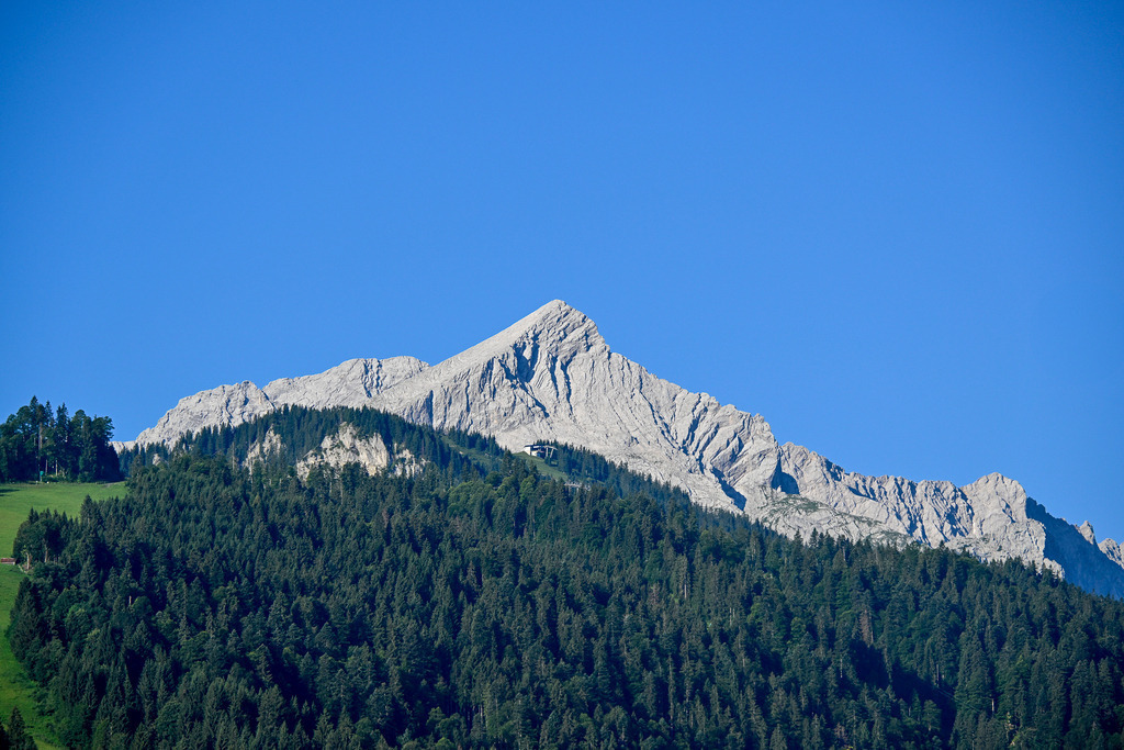 Krone der Berge: Die Alpspitze im strahlenden Sommerlicht | Ein Anblick von zeitloser Schönheit und beruhigender Beständigkeit: Stolz und unerschütterlich ragt die markante Felspyramide der Alpspitze in den tiefblauen, wolkenlosen Sommerhimmel. Die hellen Kalksteinwände zeigen ihre charakteristischen, von der Natur geformten Rinnen und Strukturen in vollkommener Klarheit. Unterhalb des schroffen Massivs erstreckt sich ein dichter Teppich aus sattgrünen Nadelwäldern, der sanft zu den Bergwiesen abfällt. Diese Aufnahme fängt die erhabene Ruhe und die majestätische Kraft des Hochgebirges ein – ein Bild, das die Sehnsucht nach Freiheit, Weite und dem Gipfelglück der Alpen spürbar macht. - Realisiert mit Pictrs.com