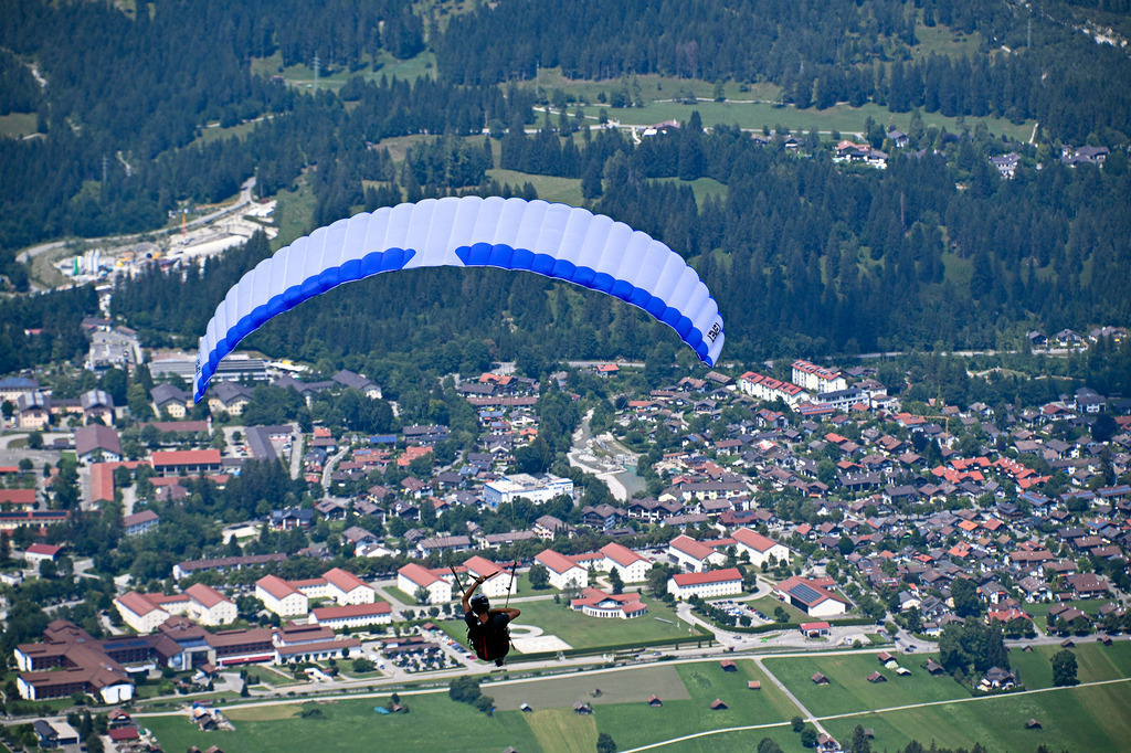Schwerelos über Garmisch-Partenkirchen | Dem Himmel so nah und die Welt zu Füßen: Ein Gleitschirmflieger schwebt majestätisch unter einem blau-weißen Schirm hoch über Garmisch-Partenkirchen. Tief unten erstreckt sich der malerische Ort, friedlich eingebettet in sattgrüne Wiesen und dichte Bergwälder. Das beeindruckende Luftbild weckt die Sehnsucht nach purer Freiheit, dem prickelnden Nervenkitzel des Fliegens und dem berauschenden Gefühl, das Zugspitzland aus der Vogelperspektive zu entdecken. - Realisiert mit Pictrs.com