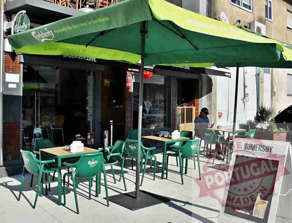 Outdoor cafe seating with green umbrellas on a sunny sidewalk | Outdoor sidewalk cafe with green Carlsberg umbrellas and tables, a man using a phone - Realized with Pictrs.com