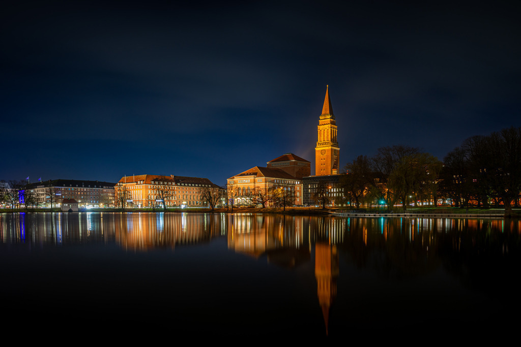 Kieler Rathaus Turm in der Nacht | Das Bild zeigt eine nächtliche Aufnahme des Kieler Rathauses mit seinem markanten Turm, der in warmem Licht beleuchtet ist. Im Vordergrund spiegelt sich das Gebäude eindrucksvoll im ruhigen Wasser des „Kleinen Kiels“. Die umgebenden Gebäude sind ebenfalls dezent erleuchtet, und die Lichter entlang der Uferpromenade erzeugen farbige Reflexionen im Wasser. Der dunkle Himmel und die ruhige Atmosphäre lassen die Architektur des Rathauses besonders zur Geltung kommen, während die Spiegelungen im Wasser die Szene harmonisch abrunden.