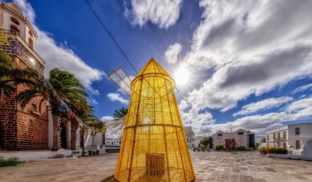 Ein Moment auf dem Platz in Costa Teguise in Lanzarote,Urlaub | Das Bild zeigt einen Moment auf der Kanareninsel Lanzarote. Ort: Der Platz befindet sich in Teguise, der ehemaligen Hauptstadt von Lanzarote.Sehenswürdigkeit: Im Hintergrund ist ein Teil der Kirche Iglesia Nuestra Señora de Guadalupe zu sehen.Objekt: Im Vordergrund ist eine große, gelbe, zeltartige Struktur zu sehen, die Teil einer temporären Installation oder Dekoration ist.Klima: Die Szene zeigt den für die Insel typischen blauen Himmel mit weißen Wolken und starkem Sonnenschein. - Realisiert mit Pictrs.com