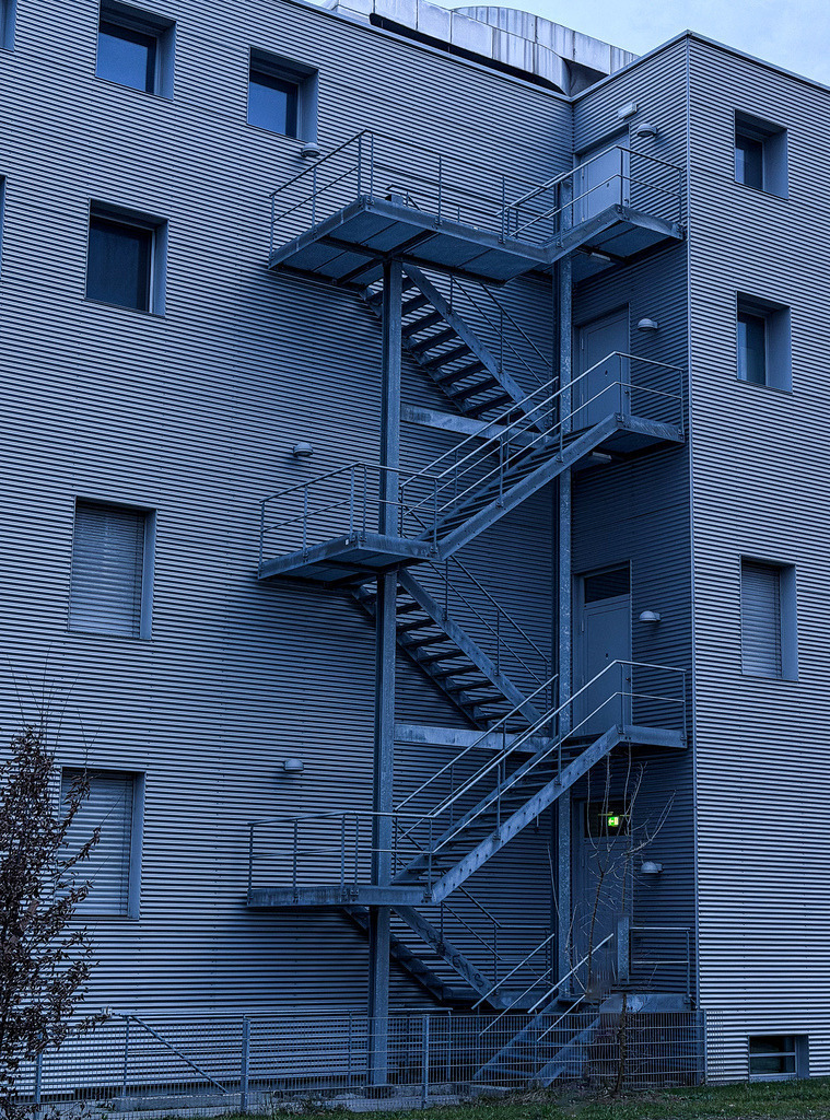 Urbane Geometrie | Moderne Architektur-Fassade Wandbild | Moderne Architektur-Fotografie mit starker grafischer Wirkung. Das kühle Blau-Grau der Industriefassade und die markante Geometrie der Außentreppe erzeugen eine faszinierende urbane Tiefe. Dieses minimalistische Kunstwerk wirkt besonders edel als großformatiger Galerie-Druck auf mattem Alu-Dibond oder hinter glänzendem Acrylglas. Perfekt für moderne Wohnräume, Lofts oder Architekturbüros im Raum Basel und Lörrach. - Realisiert mit Pictrs.com