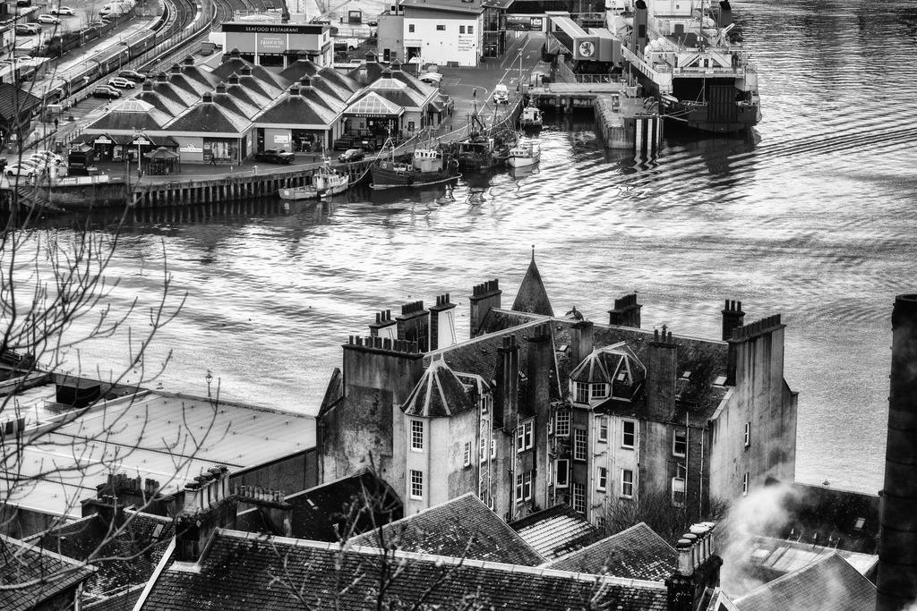 Hafenansicht Oban, Schottland | Eine erhoehte Perspektive erfasst den belebten Hafen von Oban mit Faehrterminal, Fischerbooten und angrenzendem Bahnhof. Im Vordergrund dominieren historische Gebaeude mit markanten Daechern und zahlreichen Schornsteinen, die einen Kontrast zur industriellen Hafenanlage bilden. Die Schwarz-Weiss-Darstellung hebt die strukturellen Details und die dynamischen Wasserbewegungen hervor. - Realisiert mit Pictrs.com
