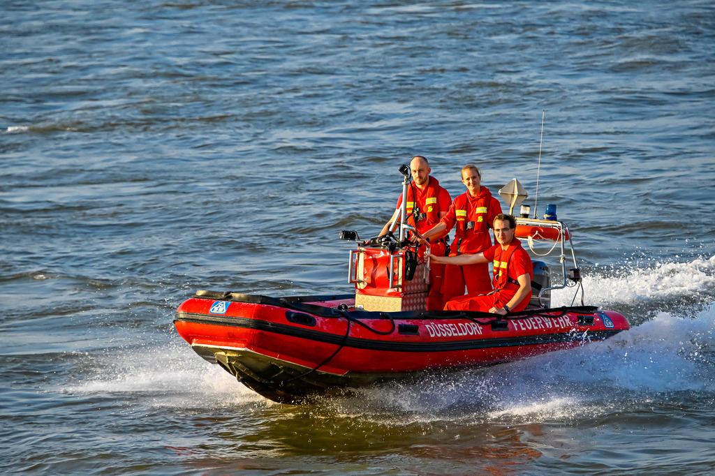 Volle Kraft voraus: Wasserrettung in Düsseldorf | Das dynamische Foto zeigt ein rotes Festrumpfschlauchboot (RIB) der Feuerwehr Düsseldorf in rasanter Fahrt auf dem Rhein. Das Boot schneidet durch das unruhige, blaugraue Wasser und erzeugt eine beeindruckende Gischtwelle am Heck. An Bord befinden sich drei Einsatzkräfte in leuchtend roter Schutzkleidung und Schwimmwesten. Die vorderen beiden stehen und bedienen technische Ausrüstung am Steuerstand, während der dritte Retter sitzt. Am Bug prangt der Schriftzug „DÜSSELDORF FEUERWEHR“. Ein starkes Bild für Professionalität und Teamarbeit im Rettungseinsatz. - Realisiert mit Pictrs.com