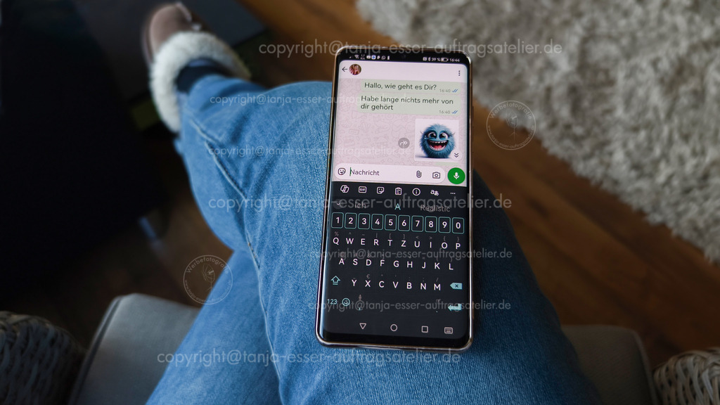 Editorial Bild zeigt einen Chat mit Whats App | Brilon, Deutschland, Februar 20ter 2026: Ein Smartphone liegt auf dem Bein einer Frau mit plüschigen Pantoffeln. Auf dem Bildschirm ist WhatsApp mit einem Chatverlauf zu sehen. Deutscher Text: „Hallo, wie geht es Dir? Habe lange nichts mehr von dir gehört“, ergänzt durch ein kleines Monster-Emoji.