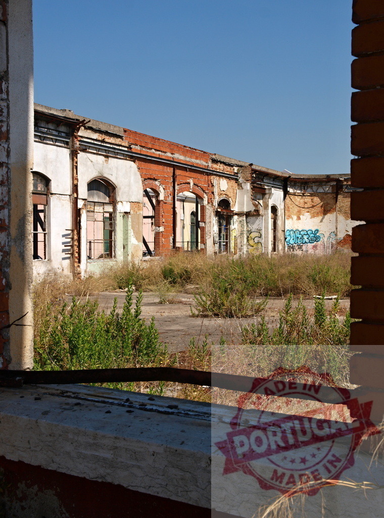 Abandoned industrial factory building decaying urban landscape view. Lisbon - Portugal | Abandoned factory view framed by a window, showing crumbling brick walls, broken windows, and overgrown weeds - Realized with Pictrs.com