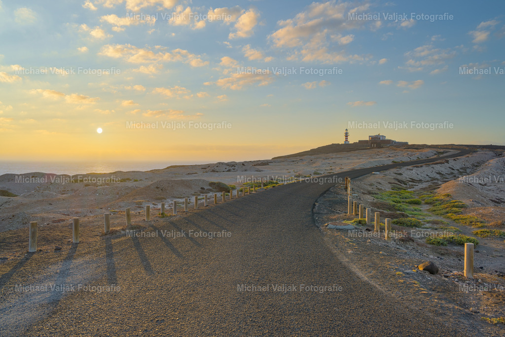 Straße zum Faro de Arinaga auf Gran Canaria | Die schmale Straße schlängelt sich durch die karge Landschaft von Gran Canaria, direkt auf den Faro de Arinaga zu. Im weichen Licht des Sonnenaufgangs wird das Bild zu einem Moment der Stille: Der Himmel ist in zarte Pastelltöne getaucht, während das goldene Licht erste Schatten auf den Asphalt wirft. Der Leuchtturm am Horizont wirkt wie ein ruhender Wächter, der dem neuen Tag entgegenblickt. - Realisiert mit Pictrs.com