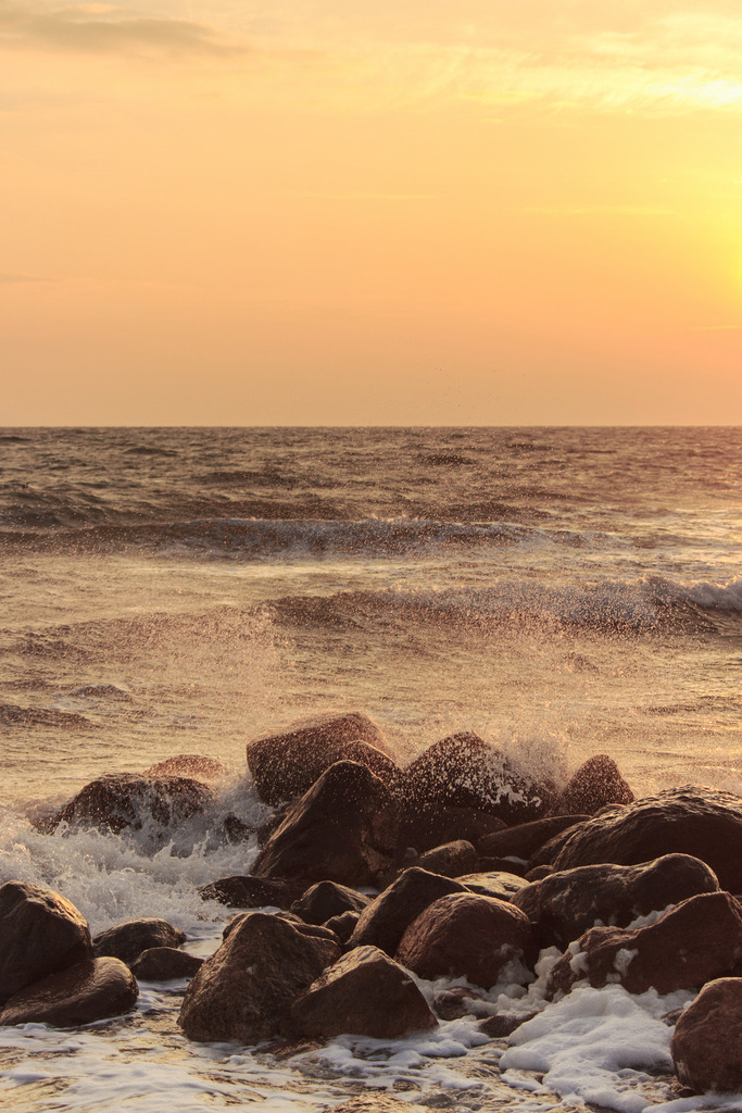 Wandbild: Morgenrot an der Ostsee | Dieses Wandbild im Hochformat zeigt einen schönen Sonnenaufgang am Meer. Der Himmel leuchtet in orangerot. Im Vordergrund sind Steine der Steinmole zu sehen. Zwischen den Steinen befindet sich der Schaum der Gischt - Realisiert mit Pictrs.com