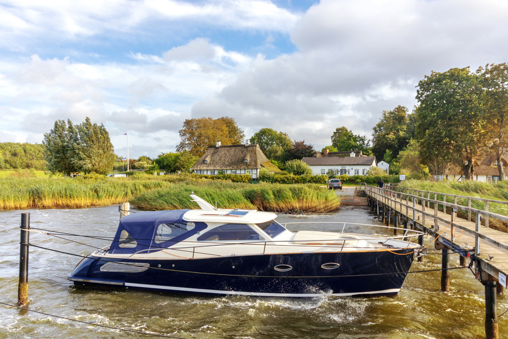 Akustikbild: Boot am Schleianleger in Sieseby an der Schlei | Dieses Akustikbild im Querformat zeigt ein Boot am Schleianleger in Sieseby zum Herbstbeginn. Auf der Schlei befinden sich kleine Wellen, die am Boot brechen. Auf der rechten Seite kann man einen Teile des Schleianlegers sehen.  Am Ufer stehen zwei malerische Häuser. Der Himmel ist überwiegend bewölkt. - Realisiert mit Pictrs.com