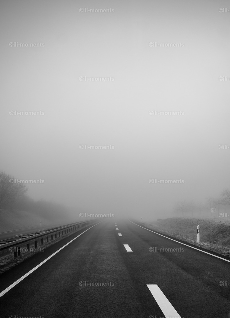 Straße im Nebel | Schwarz-weißes Foto einer leeren zweispurigen Straße im frühen Morgennebel, aufgenommen während der Fahrt. Das Bild vermittelt Ruhe und Einsamkeit – ideal als Kunstdruck für stimmungsvolle und meditative Raumgestaltungen. - Realisiert mit Pictrs.com