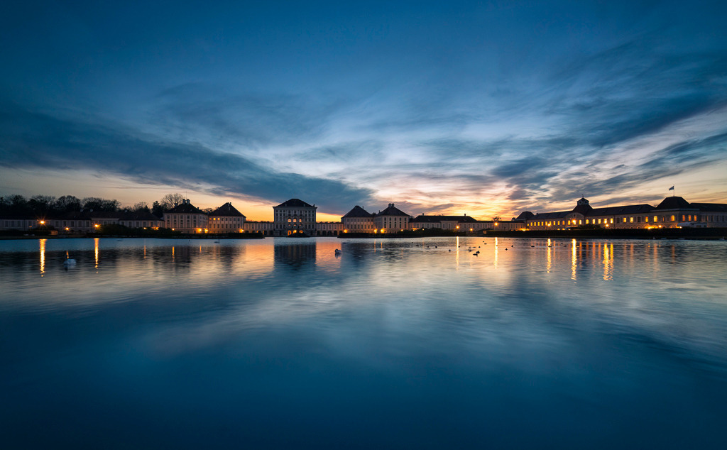 Blaue Stunde am Schloss Nymphenburg | Das Schloss Nymphenburg im sanften Licht der blauen Stunde – ein Moment voll Ruhe und Eleganz. Die Spiegelung des barocken Bauwerks im Wasser verstärkt die Symmetrie der Szene, während der Abendhimmel mit seinen letzten Farbtönen den Tag verabschiedet. Ein Bild, das München von seiner romantischsten Seite zeigt. - Realisiert mit Pictrs.com