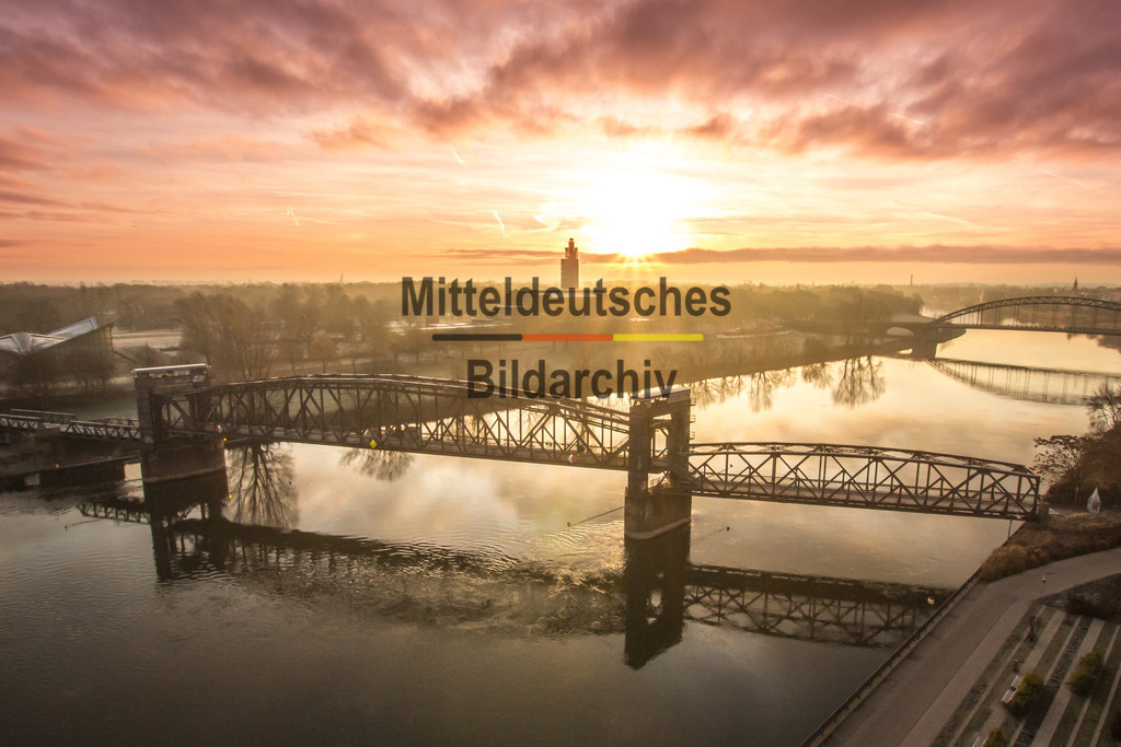 Luftbild Magdeburg Dom Hubbrücke Sonnenaufgang-0171 | Das Luftbild zeigt die historische Hubbrücke in Magdeburg während eines eindrucksvollen Sonnenaufgangs aus einer Drohnenperspektive. Im Gegenlicht zeichnet sich die markante Stahlfachwerkkonstruktion als klare Silhouette vor dem farbenintensiven Morgenhimmel ab. Dramatische Wolkenformationen verstärken die Wirkung der Aufnahme, während die Elbe das technische Bauwerk eindrucksvoll umrahmt.Die Hubbrücke gehört zu den bedeutendsten Stahlbrücken Deutschlands. Die heutige Konstruktion wurde 1933 errichtet und besitzt eine Gesamtlänge von 220 Metern. Ihr 90 Meter langer beweglicher Mittelteil mit einem Gewicht von rund 450 Tonnen konnte durch einen elektrischen Hubmechanismus um 2,87 Meter angehoben werden, um den Schiffsverkehr auf der Elbe auch bei erhöhtem Wasserstand sicher passieren zu lassen. Die genietete Stahlfachwerkkonstruktion gilt bis heute als herausragendes Beispiel des historischen Brückenbaus.Die Drohnenaufnahme verbindet die technische Präzision der historischen Stahlkonstruktion mit der eindrucksvollen Lichtstimmung eines Sonnenaufgangs. Das Zusammenspiel aus Ingenieurbau, Elbe und dramatischem Himmel macht die Hubbrücke zu einem der markantesten technischen Wahrzeichen Magdeburgs. - Realisiert mit Pictrs.com