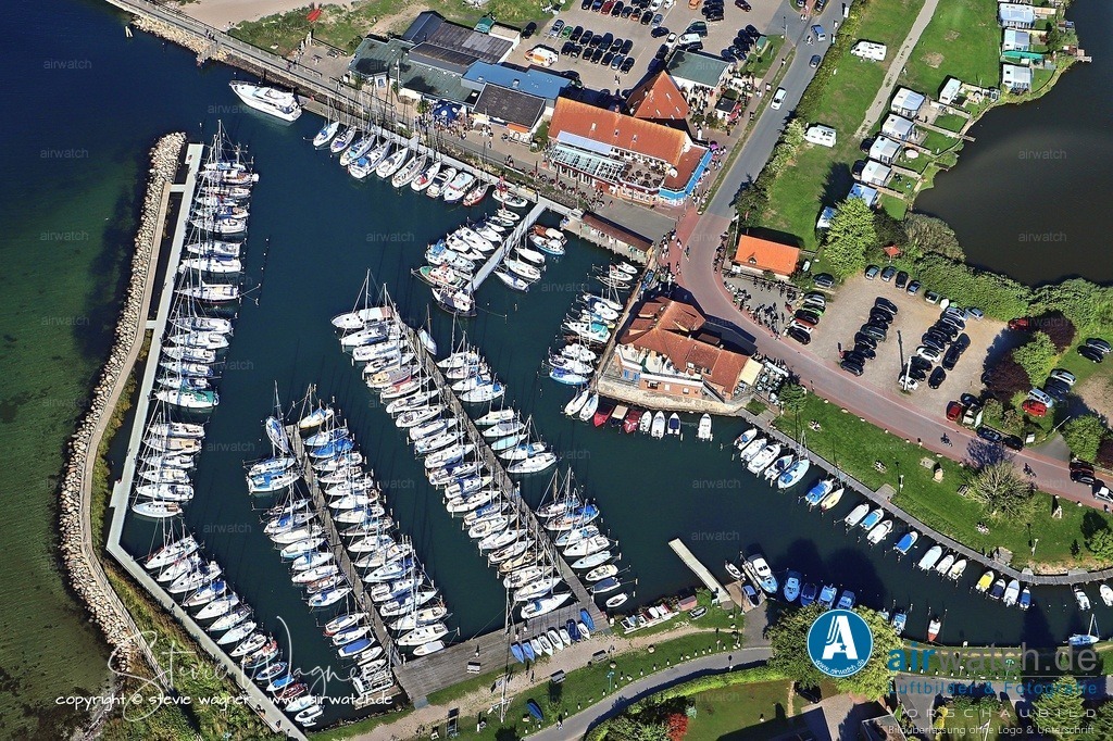 Luftbild Langballigau - Die Hafenanlage verfügt über ca. 220 Liegeplätze mit Strom- und Wasseranschluss | Die Hafenanlage verfügt über ca. 220 Liegeplätze mit Strom- und Wasseranschluss und einer Wassertiefe von 1,50 m bis 2,80 m. Die Zufahrt wurde 2019 ausgebaggert, um eine Wassertiefe von drei Metern zu erreichen, was die Anfahrt für größere Segelyachten ermöglichte. Die Hafeneinfahrt ist auch bei Dunkelheit problemlos möglich, da ein Richtfeuer (Kurs 177) vorhanden ist. Dennoch neigen die Hafeneinfahrt und der Hafen selbst zur Versandung, und ungünstige Windverhältnisse können den Wasserstand um bis zu 0,5 m senken. Freie Liegeplätze sind mit einem grünen Schild gekennzeichnet.